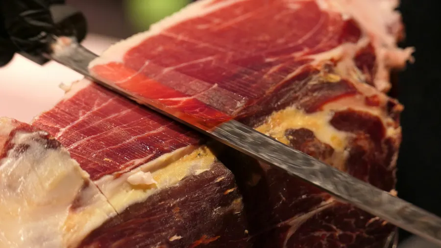 Feliere manuală de jamón iberico de bellota cu un cuțit lung special, evidențiind textura marmorată a cărnii și straturile subțiri de grăsime lucioasă.