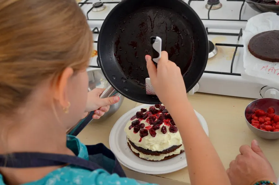 Copil la atelier de educație gastronomică, toarnă sos de fructe de pădure peste un tort cu frișcă