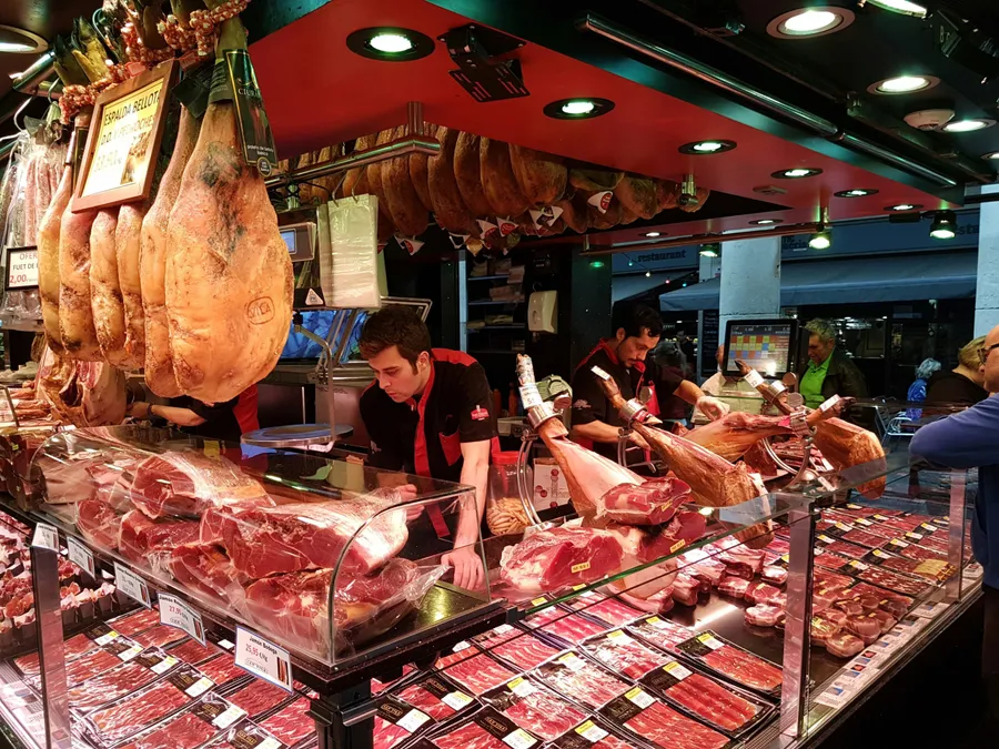 Stand tradițional dintr-o piață spaniolă, cu zeci de pulpe de jamón agățate de tavan și vitrine pline cu sortimente maturate, feliate și ambalate.